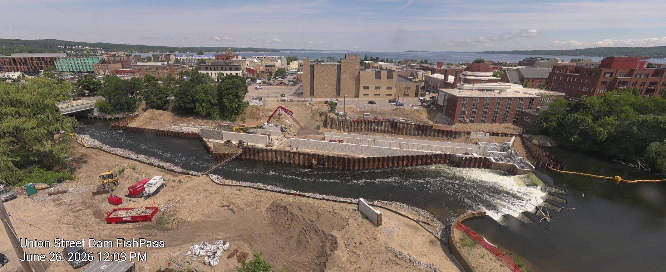 Aerial view of the Union Street Dam FishPass construction site in Traverse City, Michigan, showing a concrete channel being built alongside the river, with sheet pile walls, excavators, and downtown buildings and Grand Traverse Bay in the background. Timestamp reads June 26, 2026, 12:03 PM.
