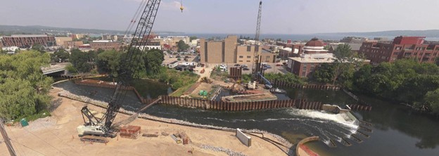 Aerial view of the Union Street Dam FishPass construction site in Traverse City, Michigan, taken in September 2025. The image shows cranes and construction equipment working along the Boardman River, with sheet piling and infrastructure forming around the developing fish passage channel. Downtown buildings and West Grand Traverse Bay are visible in the background.