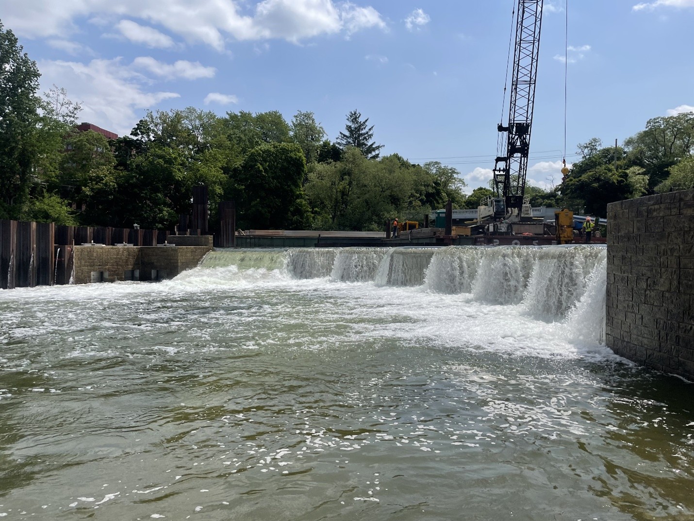 View from the river of an active construction site at a dam, where a crane and workers are present above a cascading waterfall. Concrete and steel structures frame the dam, with trees and blue sky in the background.